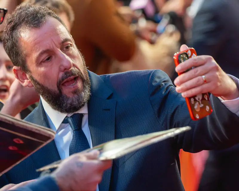 Adam Sandler in a navy suit taking a selfie with a fan's phone on a red carpet, 2025